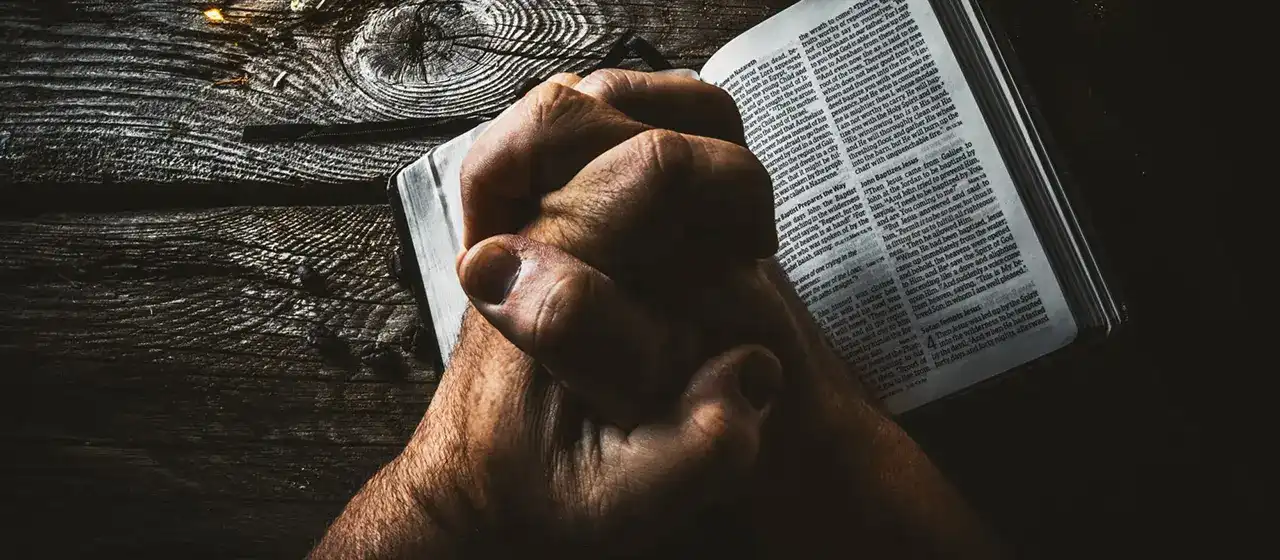 A white man's hands folded in prayer, resting on a dark wooden table with a bible.