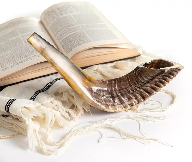 An open Bible alongside a ram's horn on a white background, with a Jewish prayer shawl.